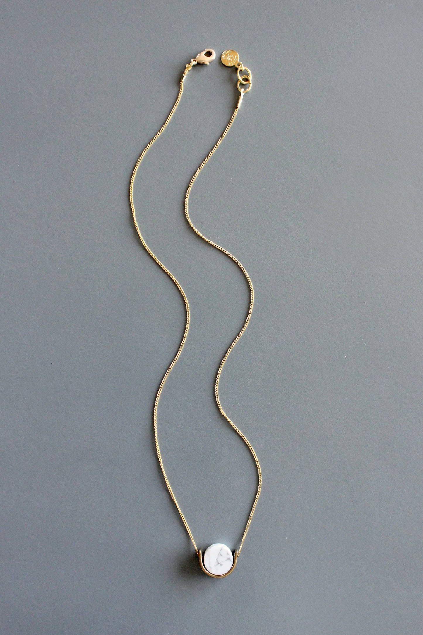 A delicate gold chain necklace with a white marble pendant by Howlite lies flat and straight down on a solid gray background.