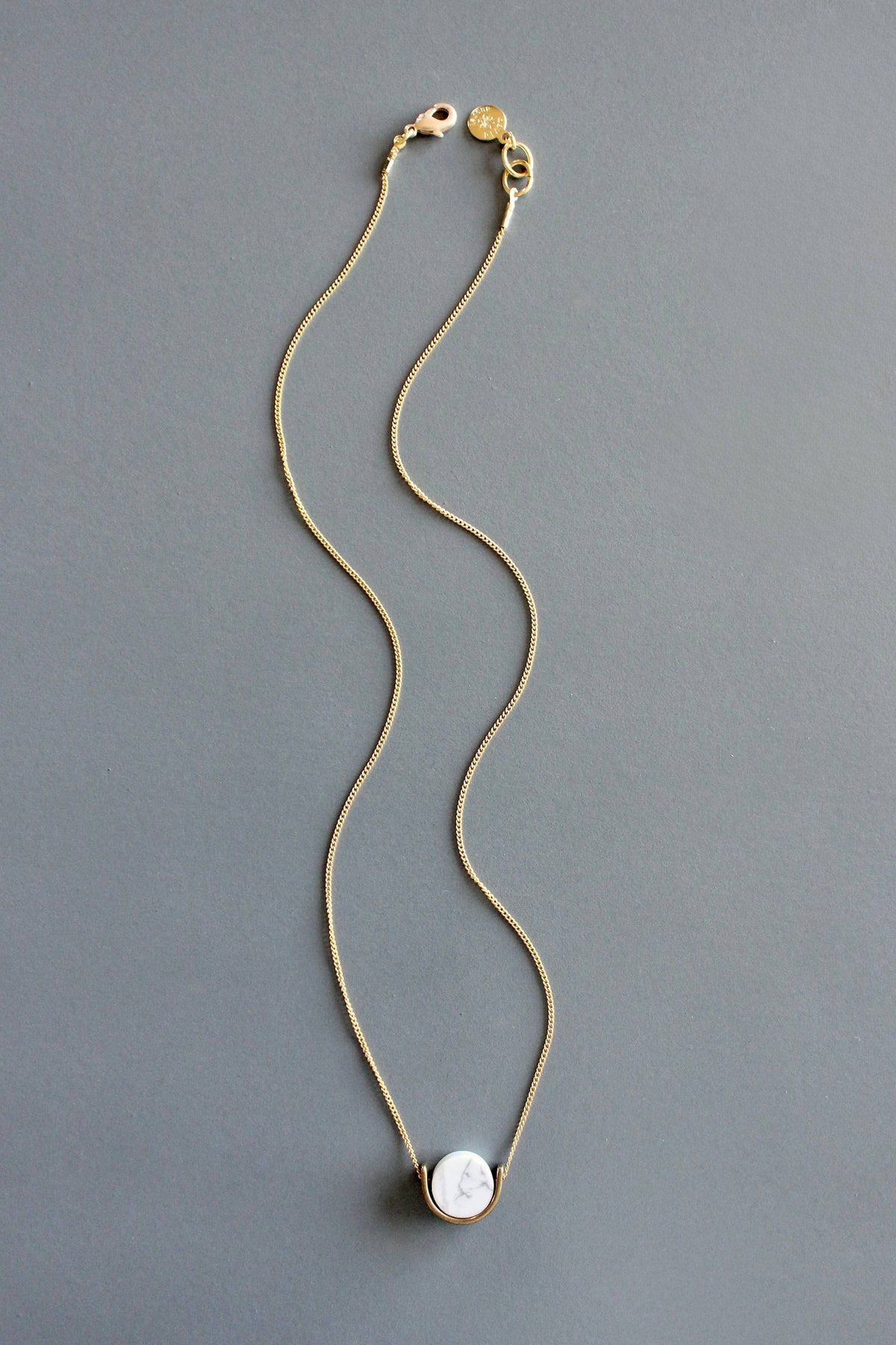 A delicate gold chain necklace with a white marble pendant by Howlite lies flat and straight down on a solid gray background.