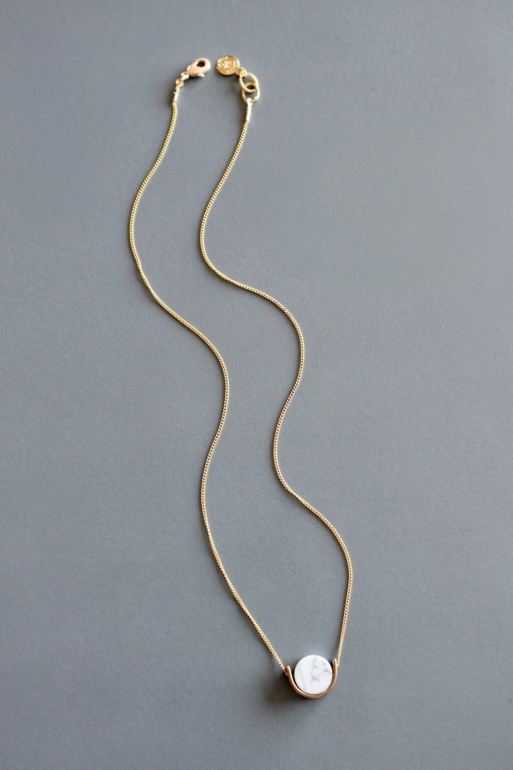 A delicate gold chain necklace with a white marble pendant by Howlite rests on a solid gray background.