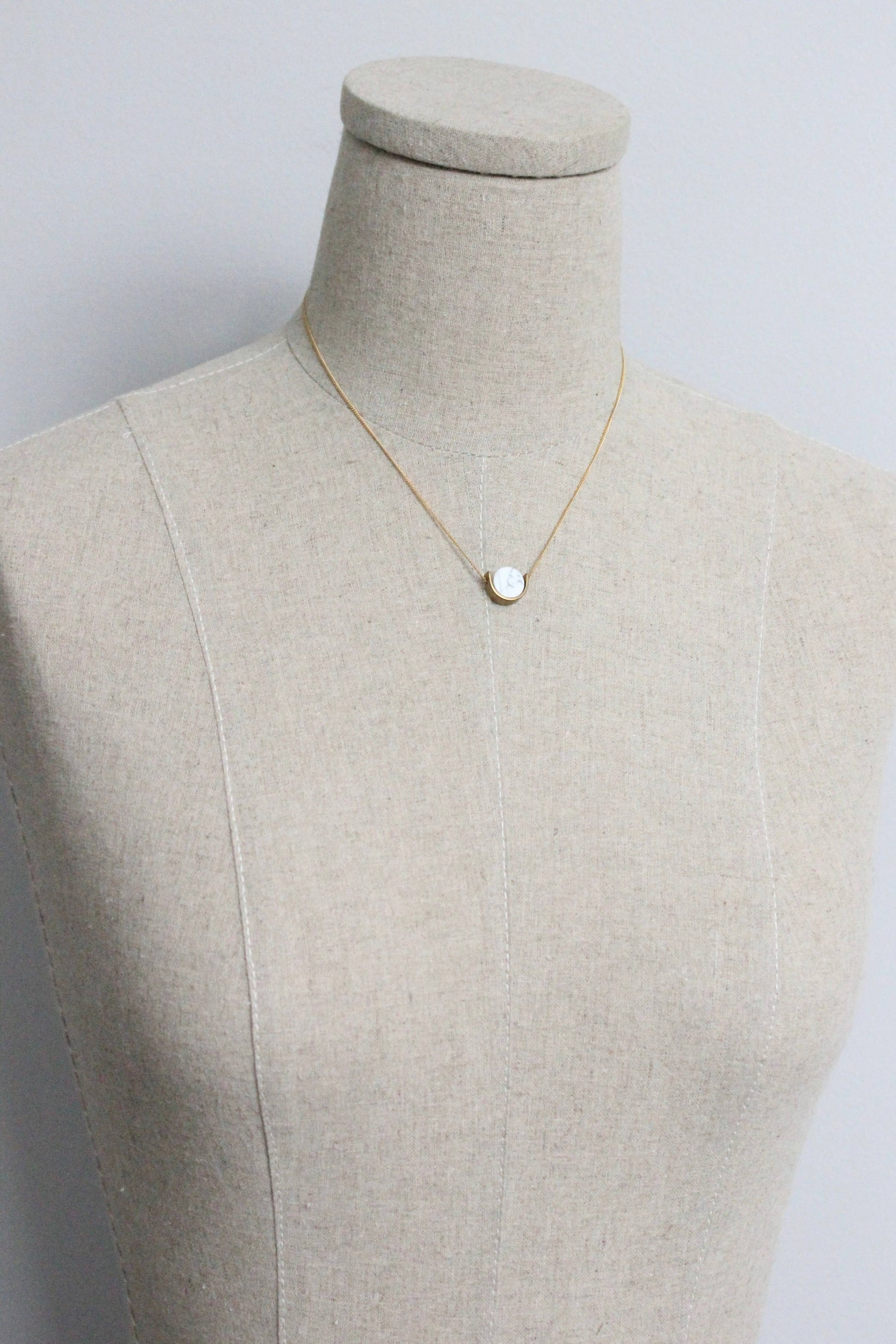 A delicate gold chain necklace with a white marble pendant by Howlite hangs from a mannequin's neck.