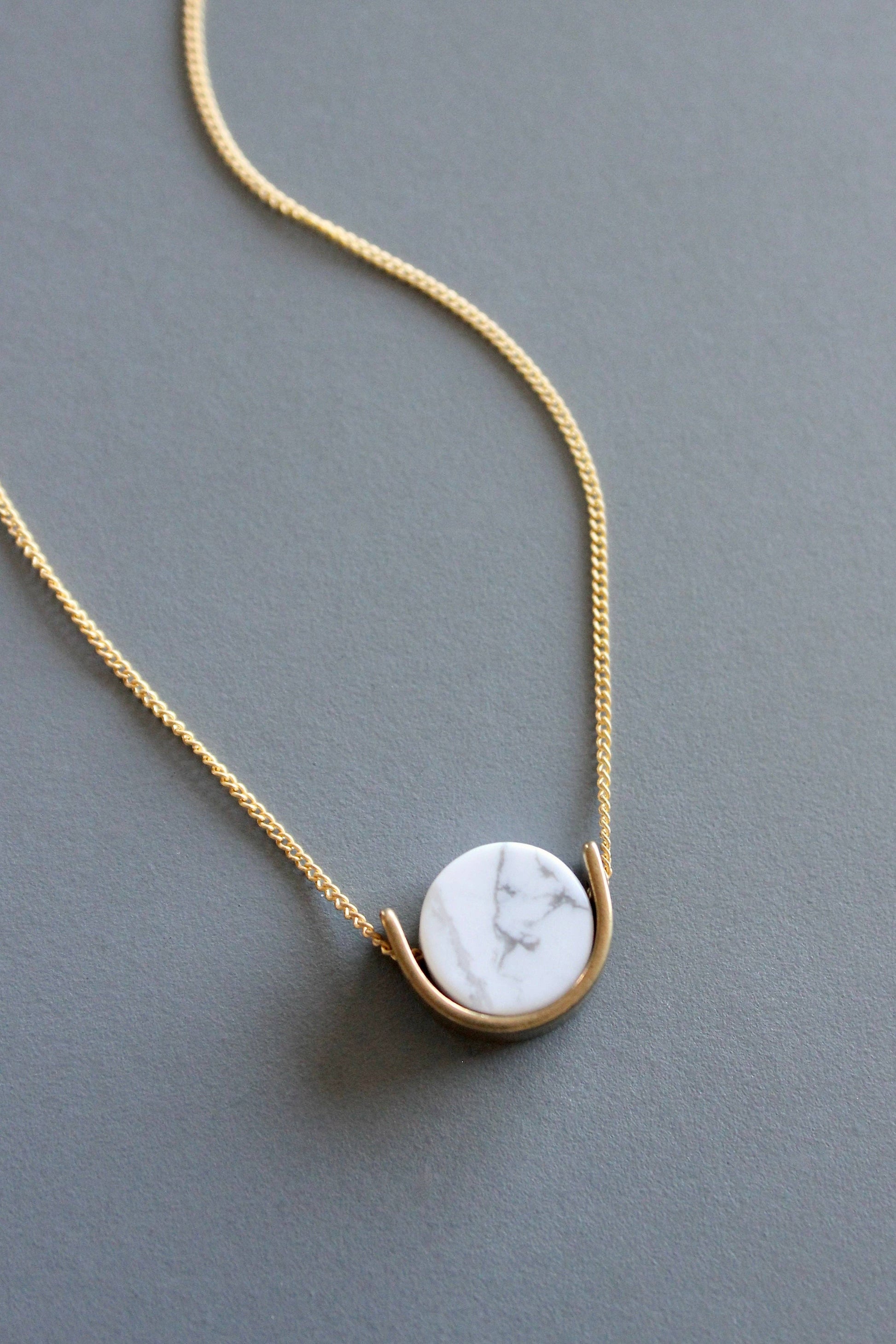 A close-up of the marble pendant on a delicate gold chain necklace by Howlite rests on a solid gray background.